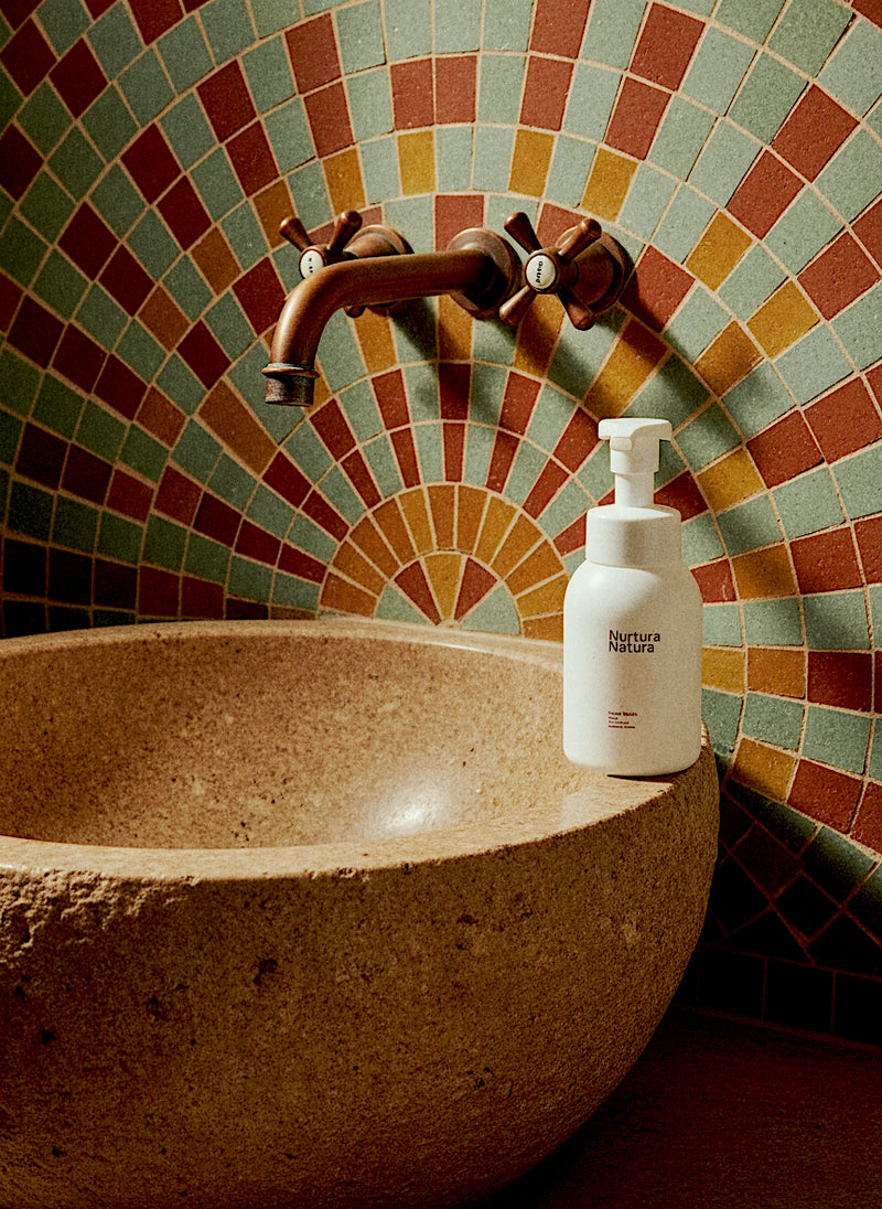 A close-up of a stone vessel sink with a white Nurtura Natura foaming dispenser bottle on its rim, set against a decorative mosaic tile backsplash with a bronze faucet.