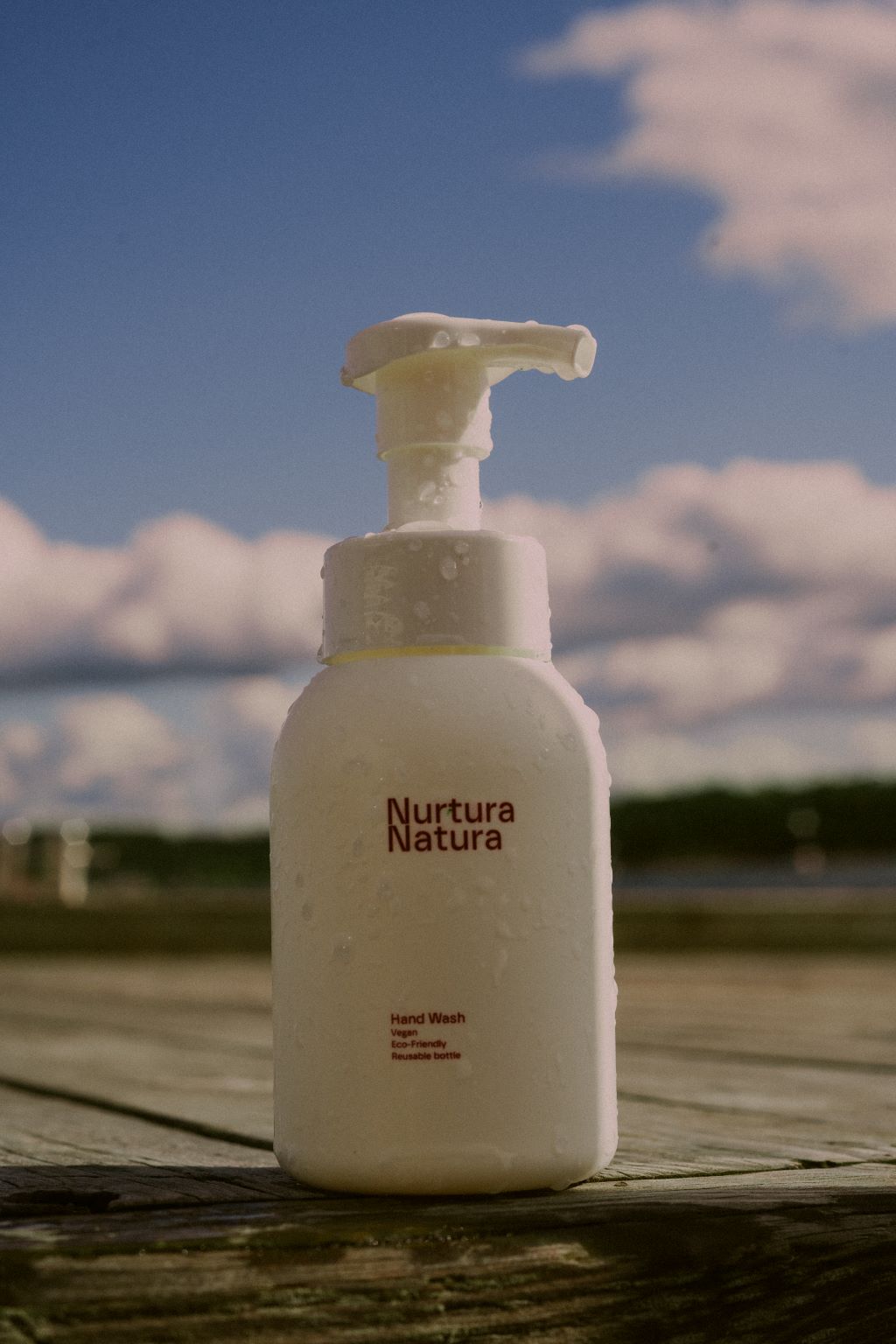 A white foaming dispenser bottle of Nurtura Natura hand wash with condensation droplets, set on a wooden deck with a blurred background of blue sky and clouds.