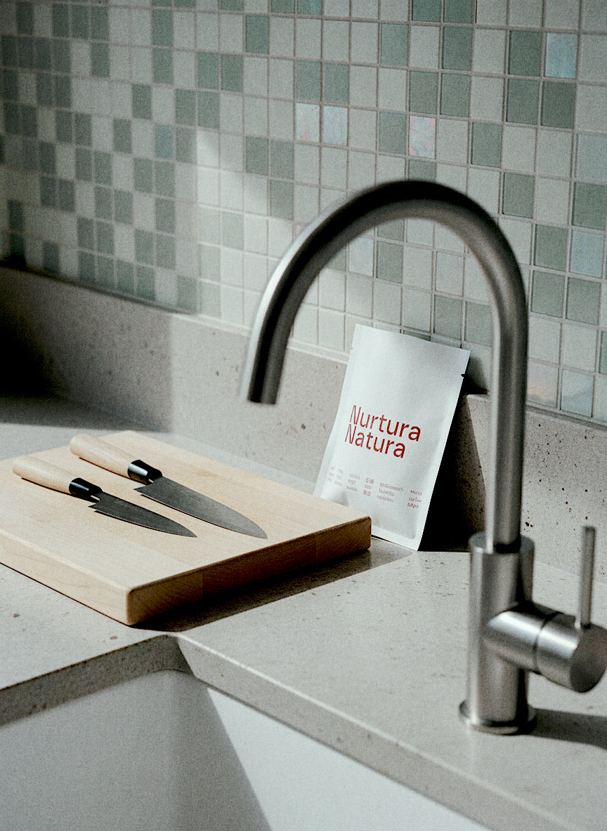 A modern kitchen scene with a tall curved stainless steel faucet, a wooden cutting board, two knives, and a Refill Pack pouch labeled 'Nurtura Natura' leaning against the backsplash.